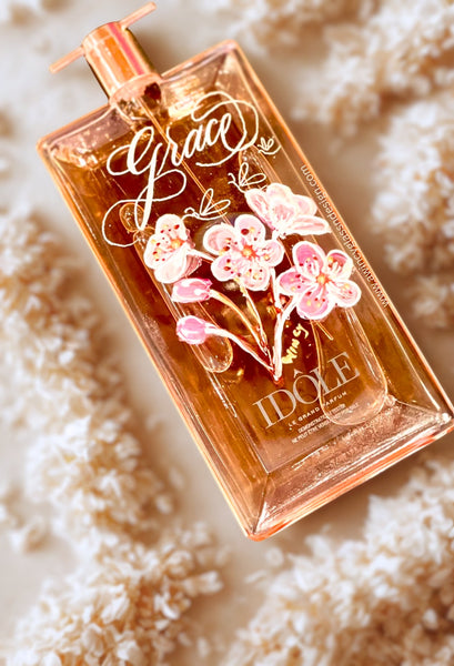Engraved and hand-painted Lancome Idôle perfume bottle with pink cherry blossom design, resting  flat lay on white flowers on a light surface — a luxury personalized bridal gift for brides, bridesmaids, and mothers of the bride