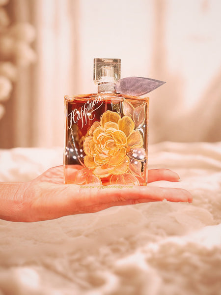 Engraved and hand-painted Lancome La Vie Est Belle perfume bottle featuring a peach peony with soft white leaves outlined in raised gold detailing. The bottle, engraved with the word ‘Happiness,’ rests elegantly in the palm of a hand lit with soft, diffused studio light against a gently blurred background and a warm champagne tone— a romantic inspired keepsake for brides, mothers of the bride, and bridal party gifting, handcrafted by A Wincy Glass ’N Desig
