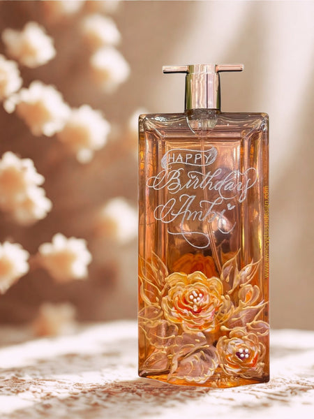Example of a personalized Lancome Idôle perfume bottle with ‘Happy Birthday’ engraving and hand-painted white and gold floral design, beside a soft pastel bouquet — crafted by A Wincy Glass ’N Design as part of our luxury engraved perfume bottle collection for brides and special occasions