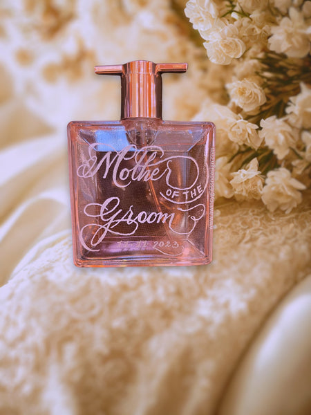 Engraved Lancôme perfume bottle reading “Mother of the Groom,” resting on a soft bridal lace with white florals; creamy champagne tones, soft vignette, shallow focus; elegant keepsake and gift idea for brides, bridesmaids, and the bridal party.