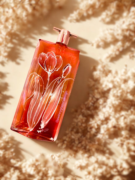 Flat lay of a Lancome perfume bottle hand-painted with pink and silver metallic tulip florals outlined in pearlescent detail,  resting  flat lay on white flowers on a light surface — a luxury personalized perfume bottle for brides, bridesmaids, and mothers of the bride, crafted by A Wincy Glass ’N Design.