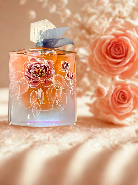 Flat lay of a Lancome La Vie Est Belle perfume bottle hand-painted with pink and silver metallic wild roses outlined in pearlescent detail, featuring raised gold accents at each bloom’s center and cascading leaves with rosebuds  — a luxury personalized perfume bottle for brides, bridesmaids, and mothers of the bride, crafted by A Wincy Glass ’N Design.